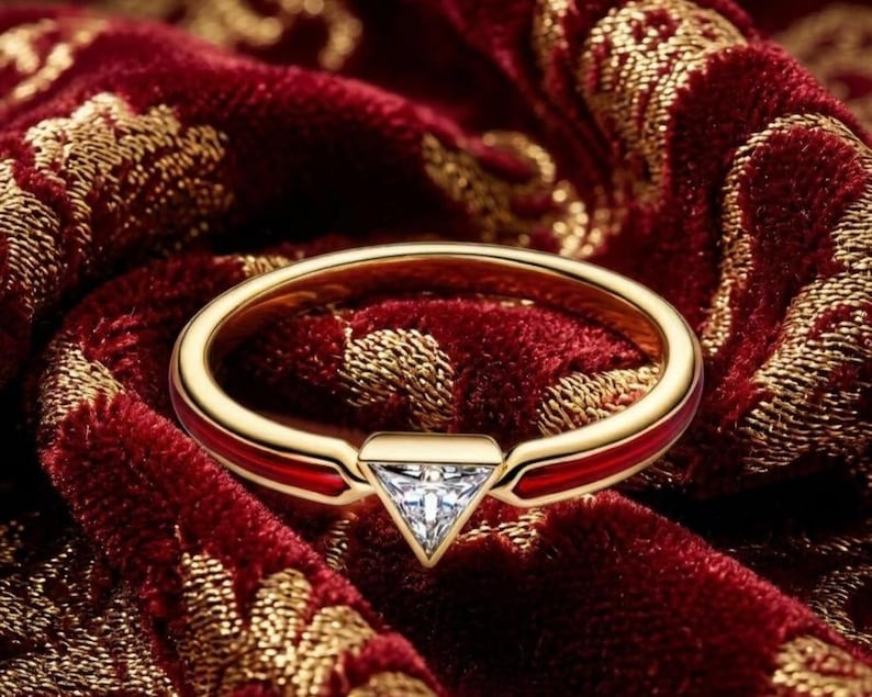 May include: A gold ring featuring a triangular-cut clear gemstone at the center. The band is accented with a smooth, deep red enamel inlay that runs along the sides, creating a striking contrast against the polished gold metal finish.