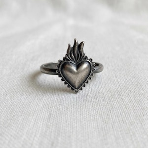 May include: A silver-toned ring featuring a heart-shaped design with a flame motif. The heart is surrounded by a scalloped edge. The ring is set against a white background.