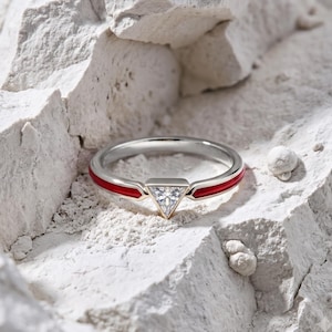 May include: A silver-toned ring featuring a central triangular-cut clear gemstone and a band accented with vibrant red enamel inlay. The ring is positioned within a textured, white, rocky surface, highlighting the contrast between the metal and stone.