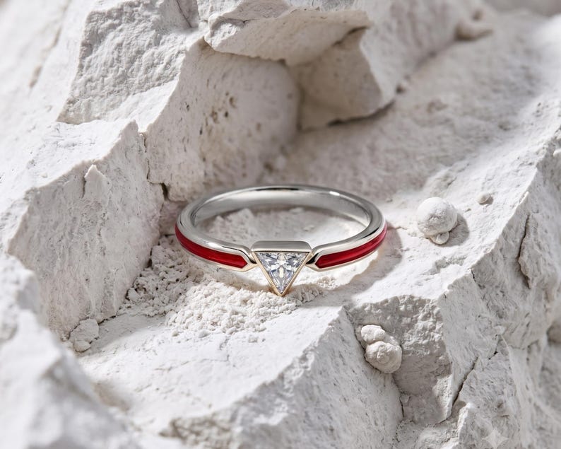 May include: A silver-toned ring featuring a central triangular-cut clear gemstone and a band accented with vibrant red enamel inlay. The ring is positioned within a textured, white, rocky surface, highlighting the contrast between the metal and stone.