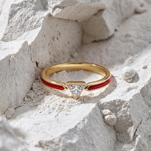 May include: A gold ring featuring a central triangular-cut clear stone set in a bezel. The band is accented with a vibrant red enamel inlay that extends from the central setting along the sides of the circular gold band, creating a sleek look.