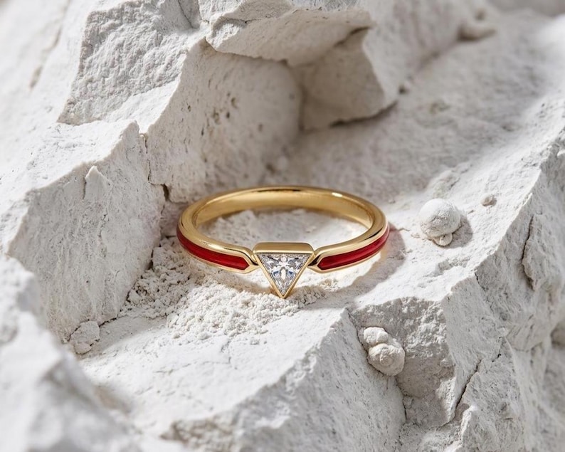 May include: A gold ring featuring a central triangular-cut clear stone set in a bezel. The band is accented with a vibrant red enamel inlay that extends from the central setting along the sides of the circular gold band, creating a sleek look.