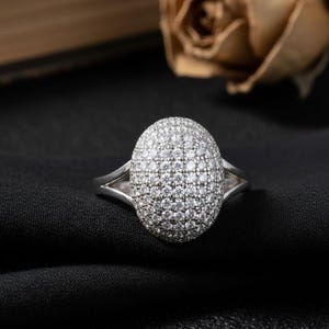 Puede incluir: Un anillo de plata con una cabeza ovalada cubierta de pequeñas piedras brillantes. El anillo está sobre una tela negra, con un fondo borroso que incluye una rosa seca.