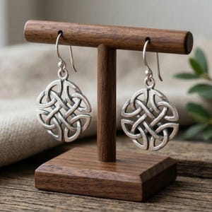 Könnte beinhalten: Ein Paar silberne Ohrringe mit keltischem Knotenmuster, präsentiert auf einem Holzschmuckständer. Die Ohrringe haben ein kompliziertes, ineinander verwobenes Design. Der Ständer ist aus dunklem Holz.