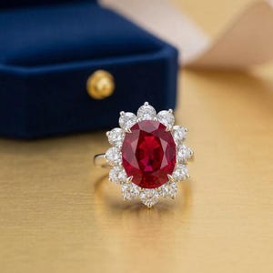May include: A silver ring with a large, oval ruby gemstone encircled by smaller, round diamonds. The ring is set on a gold surface, with a blue velvet ring box visible in the background.