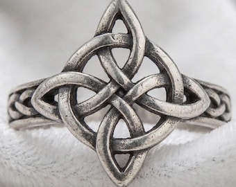 May include: A silver-toned ring featuring a central four-pointed knot design with an intricate, continuous interlaced pattern. The metal has a darkened, oxidized finish in the recessed areas, highlighting the woven structure of the band.