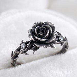 May include: A silver ring with a rose design. The ring has a thorny band with two small, clear gemstones. The rose is the focal point, with detailed petal work. The ring is set against a soft, white background.
