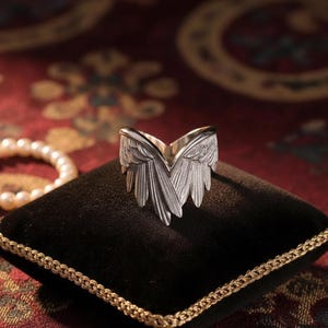 Op de afbeelding: Een zilverkleurige ring in de vorm van engelen vleugels, gepresenteerd op een zwart fluwelen kussen met een gouden rand. Een parelketting is op de achtergrond te zien. Het ontwerp van de ring heeft gedetailleerde veertjes, wat een opvallend sieraad creëert.