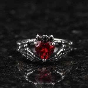 May include: A silver ring with a heart-shaped, faceted red gemstone. The ring's design includes skeletal hand details and small black gemstones at the top, creating a gothic aesthetic. The ring is a close-up shot.