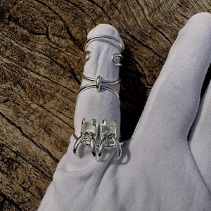 May include: A close-up of several silver rings. One ring features two square-cut stones, while others have a minimalist, wire-like design. The rings are displayed on a white fabric, set against a natural wood background.