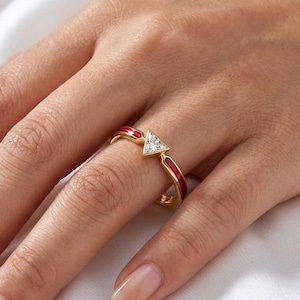 May include: A gold ring featuring a central triangular-cut clear gemstone. The band is accented with vibrant red enamel inlay on both sides of the stone, creating a distinct contrast against the polished gold metal finish of the ring structure.