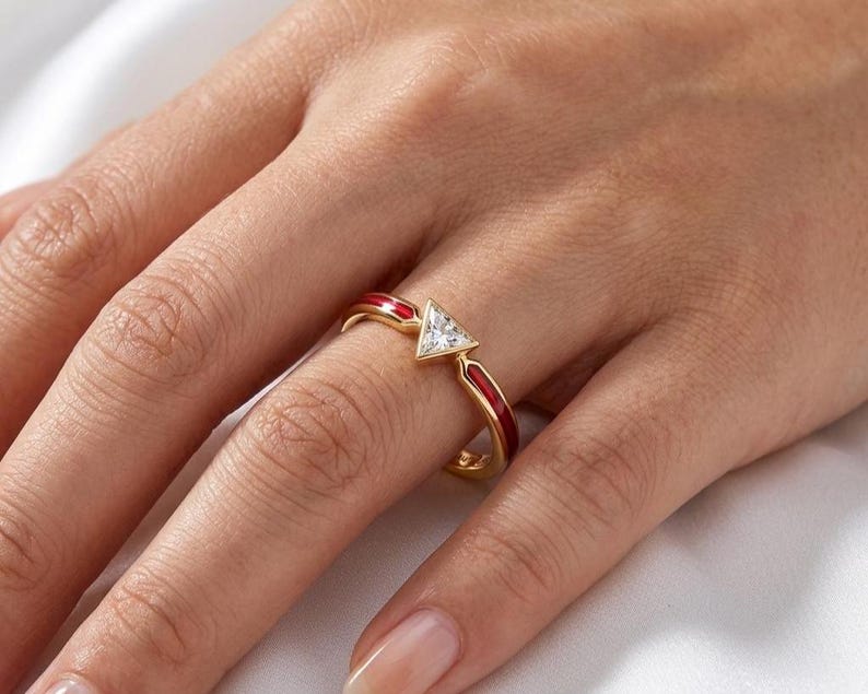 May include: A gold ring featuring a central triangular-cut clear gemstone. The band is accented with vibrant red enamel inlay on both sides of the stone, creating a distinct contrast against the polished gold metal finish of the ring structure.