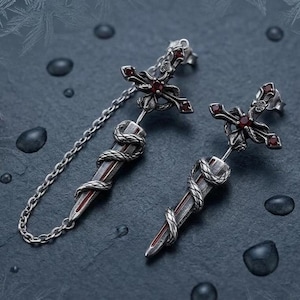 Könnte beinhalten: Ein Paar silberfarbene Ohrringe mit Kreuzdesign, roten Akzenten und einem dolchförmigen Anhänger. Der Dolch ist mit einer Schlange umwickelt und mit einer Kette mit dem Kreuz verbunden. Die Ohrringe sind vor dunklem Hintergrund.
