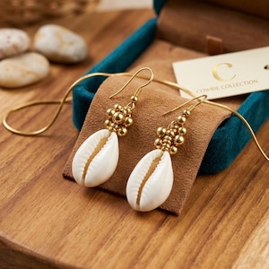 May include: A pair of gold-colored earrings featuring white seashell pendants. The earrings have a cluster of small gold beads above the shell and a gold hook. The earrings are displayed on a brown surface inside a teal box.