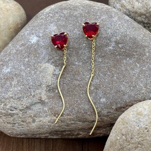 May include: A pair of gold-coloured earrings with heart-shaped, red gemstones. The earrings have a chain and a curved, gold-coloured wire that dangles below the heart-shaped stone. The earrings are displayed on a grey stone surface.