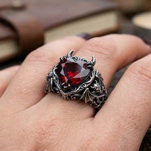Może przedstawiać: Srebrny pierścionek z dużym, fasetowanym, czerwonym kamieniem szlachetnym w kształcie serca. Pierścionek ma skomplikowane detale, w tym małe rogi i skrzydła nietoperza. Pierścionek jest na palcu.