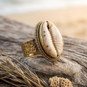 Puede incluir: Un anillo de latón con una pieza central de concha grande, ovalada y de color crema. El anillo tiene una banda texturizada y un borde en forma de cuerda alrededor de la concha. El anillo está sobre una superficie de madera envejecida.