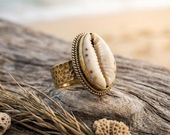 May include: A brass ring with a large, oval, cream-colored seashell centerpiece. The ring has a textured band and a rope-like border around the shell. The ring is set against a weathered wooden surface.