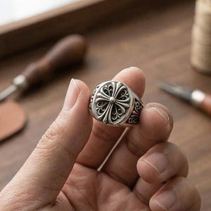 Puede incluir: Un anillo plateado con un diseño de cruz ornamentado. El anillo tiene una superficie detallada y texturizada y se sostiene en una mano. El diseño del anillo presenta un motivo de cruz central rodeado de intrincados patrones. El anillo es una joya.