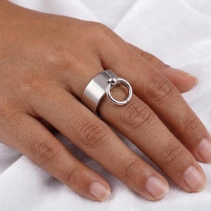 May include: A silver ring with a thick band and a small ball connected to a larger ring. The ring is worn on a finger, showcasing its modern design. The ring's polished surface reflects light, highlighting its sleek, minimalist style.