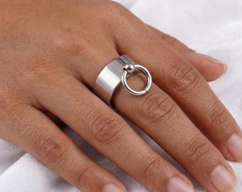 May include: A silver ring with a thick band and a small ball connected to a larger ring. The ring is worn on a finger, showcasing its modern design. The ring's polished surface reflects light, highlighting its sleek, minimalist style.