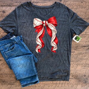 May include: A dark gray V-neck t-shirt with a baseball-themed bow design. The bow features red and white ribbons with baseball stitching details. Blue denim jeans are folded next to the shirt.