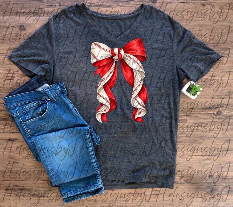 May include: A dark gray V-neck t-shirt with a baseball-themed bow design. The bow features red and white ribbons with baseball stitching details. Blue denim jeans are folded next to the shirt.