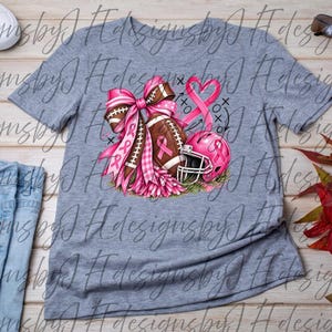 May include: A heather gray t-shirt featuring a graphic print of a brown football, a pink helmet, and a large pink ribbon bow. The design includes a pink awareness ribbon and black tic-tac-toe markings. The shirt is displayed with white sneakers, sunglasses, a red bracelet, and blue jeans.