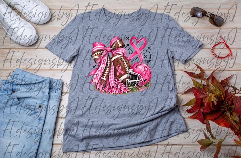 May include: A heather gray t-shirt featuring a graphic print of a brown football, a pink helmet, and a large pink ribbon bow. The design includes a pink awareness ribbon and black tic-tac-toe markings. The shirt is displayed with white sneakers, sunglasses, a red bracelet, and blue jeans.