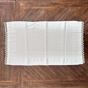 Könnte beinhalten: Ein rechteckiger, cremefarbener Tischläufer aus Leinen mit zarten Spitzenborten an den Rändern. Der Läufer liegt flach auf einer dunklen Holzoberfläche und zeigt seine schlichte Eleganz und Textur. Die Spitze verleiht einen Hauch von Vintage-Charme.