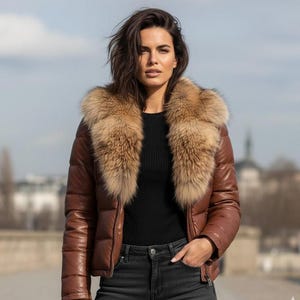 Puede incluir: Chaqueta de cuero marrón con cuello de piel. La chaqueta tiene un diseño acolchado y cierre de cremallera. El cuello de piel es de color marrón claro. La persona lleva una camiseta negra y vaqueros negros.