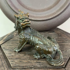 May include: A bronze-colored figurine of a mythical creature, possibly a dragon or lion, with intricate details. The sculpture has a textured surface and is posed in a seated position. The figurine is displayed on a wooden surface.