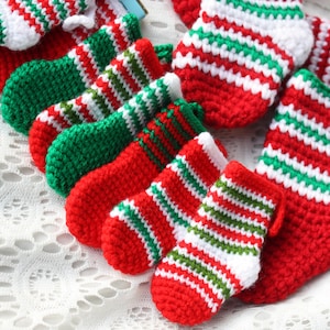 May include: A group of seven small, crocheted Christmas stockings in red, white, and green stripes. The stockings are arranged in a row on a white lace tablecloth.