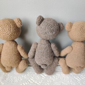 Pu&ograve; includere: Tre orsacchiotti di peluche all'uncinetto, uno marrone, uno marrone chiaro e uno grigio, sono seduti su una superficie di uncinetto bianca. Gli orsacchiotti sono rivolti con le spalle alla telecamera, mostrando la schiena.