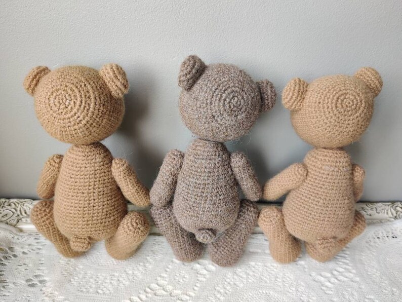 Pu&ograve; includere: Tre orsacchiotti di peluche all'uncinetto, uno marrone, uno marrone chiaro e uno grigio, sono seduti su una superficie di uncinetto bianca. Gli orsacchiotti sono rivolti con le spalle alla telecamera, mostrando la schiena.