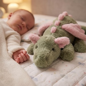 May include: A green plush dragon toy with pink wings and ear details lies on a patterned blanket. A sleeping baby is next to the toy. The soft toy is designed for comfort and companionship.