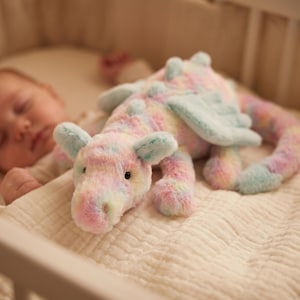 May include: A pastel-coloured plush dragon toy in pink, blue, and yellow. The dragon has small wings and ears, and is lying on a cream-coloured blanket. The toy is in a cot.