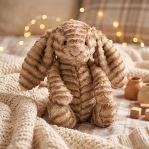 May include: A plush bunny toy in shades of brown and cream, with a striped pattern. The bunny has long, floppy ears and a pink nose. It is sitting on a cream-colored knitted blanket. The background includes soft lighting and decorative elements.