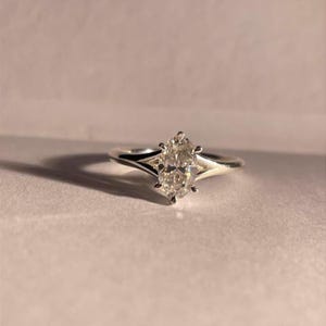 Op de afbeelding: Een zilveren verlovingsring met een ovaalvormige diamant. De diamant is gezet met zes klauwen en de band heeft een gespleten ontwerp. De ring wordt gepresenteerd op een lichtgekleurd oppervlak.