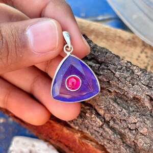 Peut inclure: Un pendentif triangulaire composé d'une pierre facettée violet foncé sertie dans une monture en métal argenté. Une petite pierre circulaire rose vif est incrustée au centre de la pierre violette, avec une bélière en métal argenté au sommet.