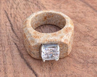 May include: A unique ring featuring a light brown, textured band with a square, clear gemstone set in a silver metal. The ring is made of a natural material, possibly coral or stone, and sits on a wooden surface.