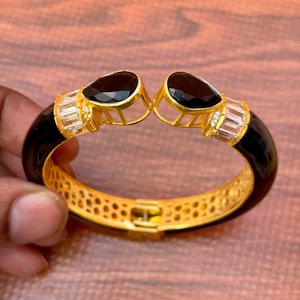 Puede incluir: Un brazalete rígido articulado en tono dorado con una banda negra lisa, dos piedras negras en forma de pera en la abertura y piedras transparentes de corte baguette en los laterales. El interior de la banda de metal dorado tiene un patrón de panal.