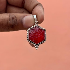 May include: A hexagonal, red gemstone pendant with a floral design carved into its surface. The pendant is set in a silver-colored metal frame with a beaded border and a small loop for a chain. The gemstone is held by a hand against a peach background.