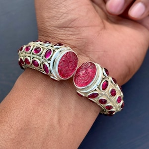 Puede incluir: Brazalete articulado de plata con piedras preciosas de rubí ovaladas y diseño floral tallado. El brazalete presenta una serie de rubíes ovalados engastados en una banda de plata, con un gran rubí tallado en el cierre.