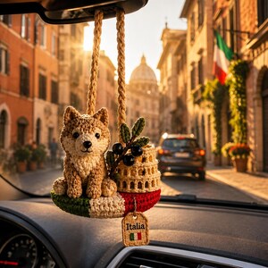 May include: A crocheted car hanging ornament featuring a tan and white dog sitting on a base colored with green, white, and red stripes. It includes a small crocheted Colosseum, a sprig of black olives, and a rectangular tag labeled Italia.