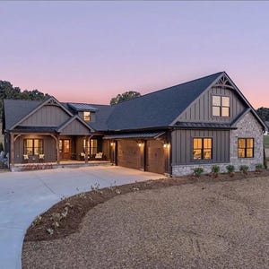 May include: Exterior view of a large, modern farmhouse with a dark gray roof and siding. The house features stone accents, multiple windows, and a two-car garage. A concrete driveway leads to the house, and the sky is a soft pink and purple.