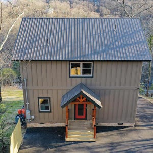 以下が含まれることがあります： グレーの金属屋根と茶色の外壁の2階建ての家。玄関ドアは赤く、ポーチには木製の支柱があります。両方の階に窓が見えます。家は木々と舗装された私道に囲まれています。