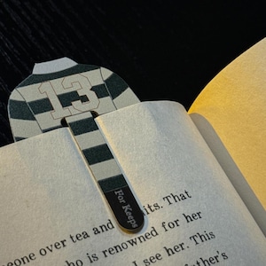 May include: A bookmark shaped like a rugby jersey with the number 13, in green and white stripes. The bookmark is inserted into a book, with the text "For Keeps" printed on the bottom. The book's pages are visible.