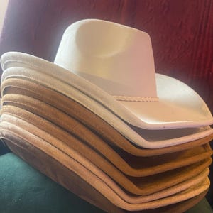 Può includere: Una pila di cappelli da cowboy in varie tonalità di marrone e beige. Il cappello superiore è di colore crema chiaro con una fascia intrecciata. I cappelli sono impilati, mostrando i loro diversi colori e trame.