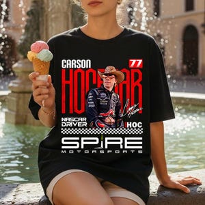 May include: A black t-shirt featuring a graphic print of NASCAR driver Carson Hocevar wearing a cowboy hat. The design includes the text Carson Hocevar, the number 77, Spire Motorsports, and a checkered flag pattern in red, white, and black.